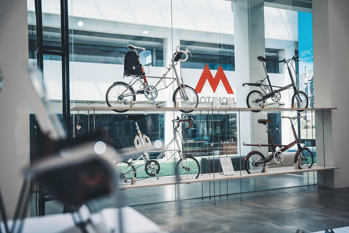 Four Moulton bikes suspended in front of large glass window with Moulton logo behind.