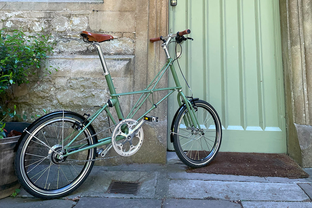 Green Moulton bike leaning against an old light green wooden door and on a paved stone floor.